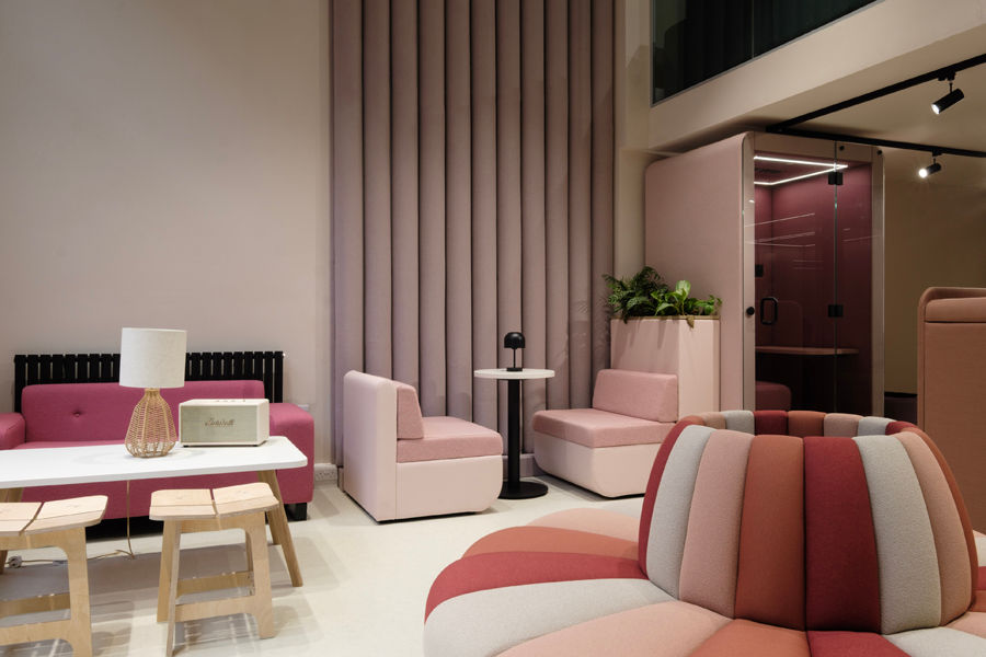 Modern lounge with pink and red seating, a white table, lamp, stools, and plant. Curtains and enclosed booth in background create a cozy vibe.
