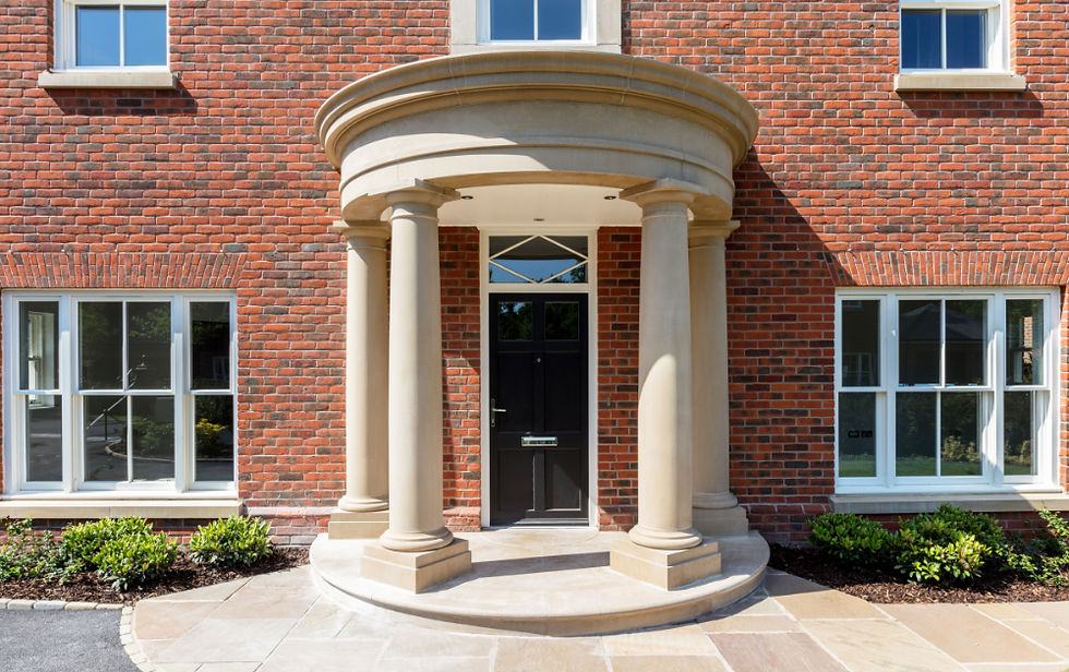 Brick building entrance with cream columns, black door, and white-framed windows in bright sunlight.