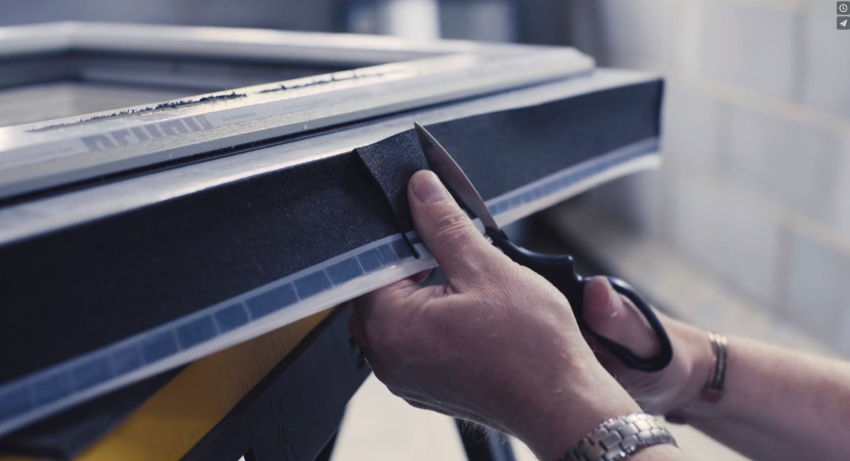 Hands use scissors to trim black material on a machine in a workshop, with a focused, hands-on feel.