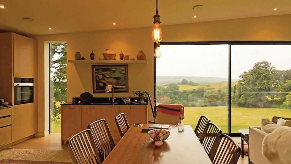 Modern dining room with wooden table, chairs, hanging lights, and large window showing a green landscape. Warm, cozy atmosphere.