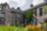 Stone cottage with ivy and teal windows, colorful wildflowers in front, beneath a cloudy sky.