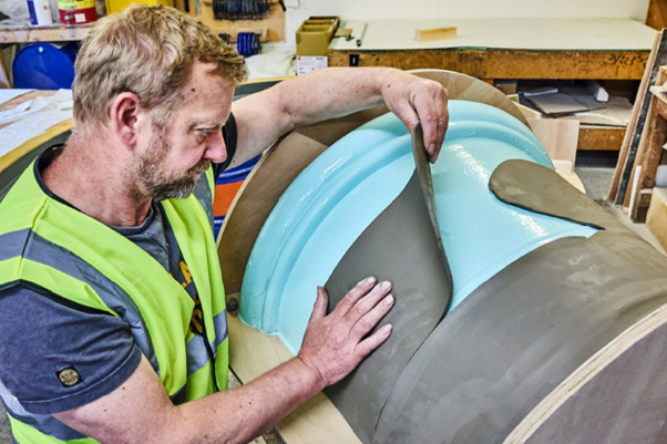 Man in a yellow vest works on sculpting a large clay object with light blue accents in a workshop setting, focused and precise.