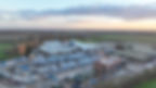 Aerial view of an industrial site with warehouses, parked trucks, and numerous shipping containers. Fields and a sunset in the background.