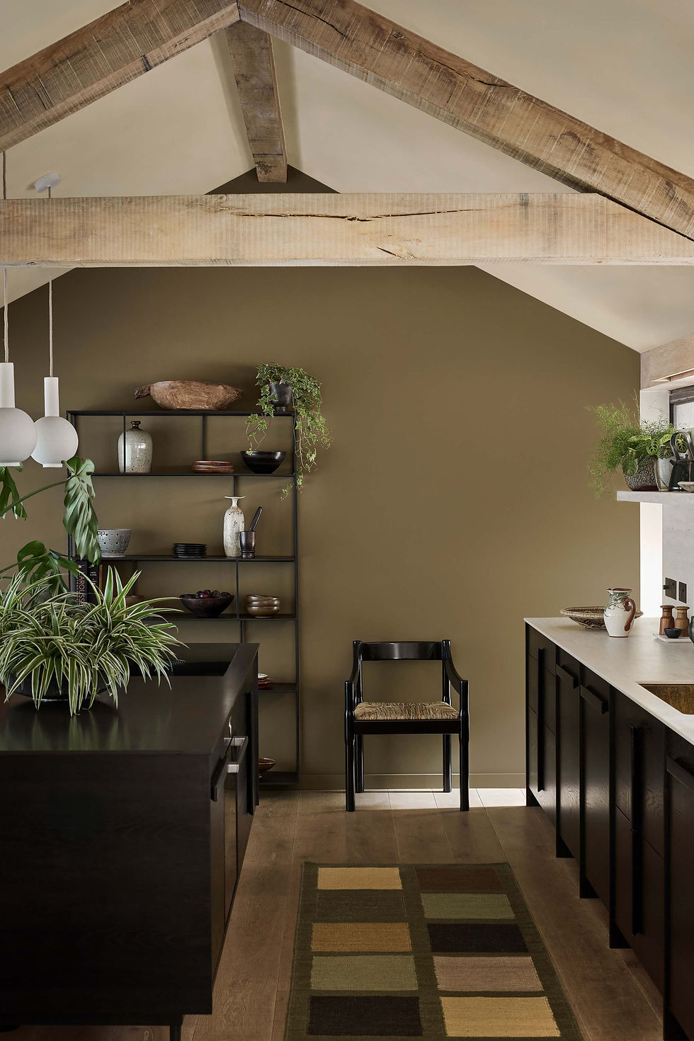 Modern kitchen with dark cabinets, open shelving, and potted plants. Wooden beams, earthy wall color, and a patterned rug create a warm ambiance.