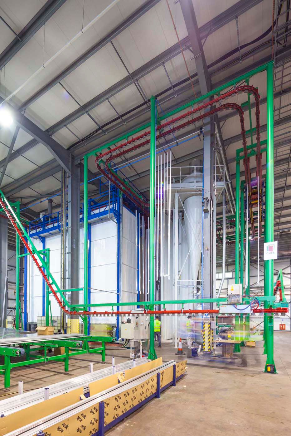 Industrial warehouse with green and red machinery, conveyor belt, and large pipes. Bright lighting and reflective surfaces. Safety cones visible.