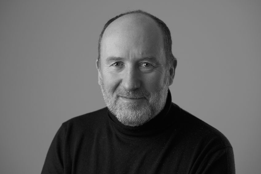 Black-and-white portrait of a smiling bald, bearded man in a black turtleneck against a plain gray background.