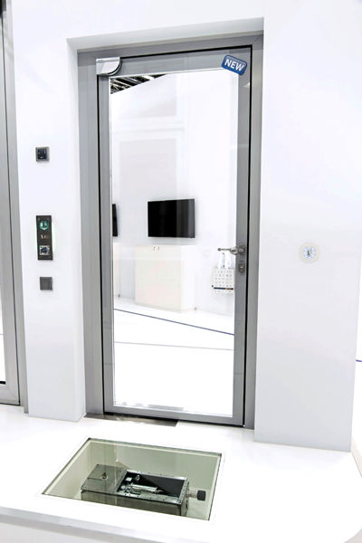 Glass door with a "NEW" label in a modern, white room. Floor has a small square glass panel revealing mechanical parts beneath. Minimalist decor.