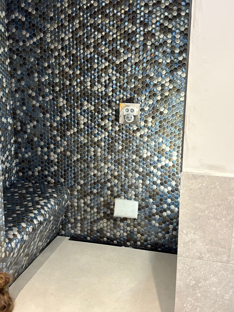 Hexagonal mosaic tiles in blue and gray adorn a wall with a square fitting, adjacent to beige tiles and a cream floor.