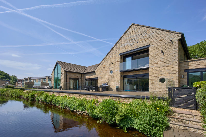 Modern stone house with glass balcony beside a pond, landscaped greenery, and a bright blue sky with contrails.