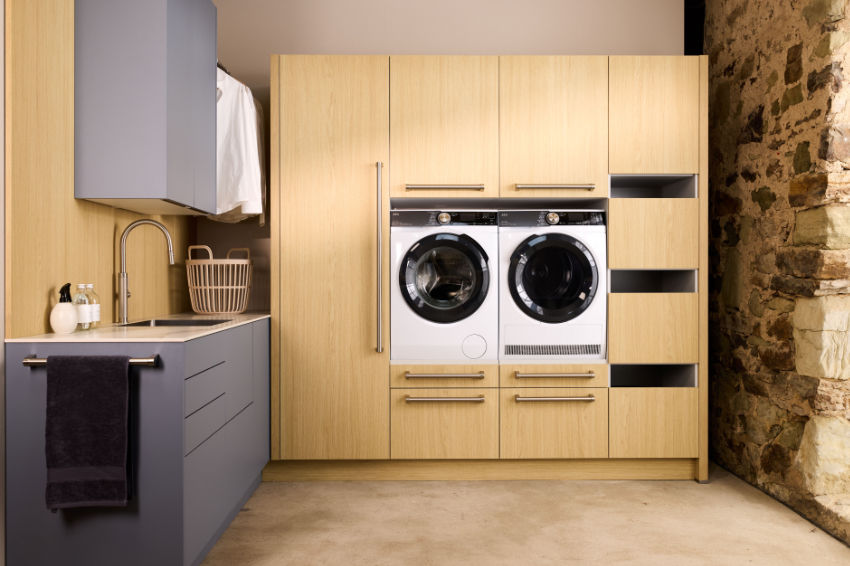Modern laundry room with wood cabinets, front-load washer and dryer, sink, and stone wall; dark towel hangs on gray counter.