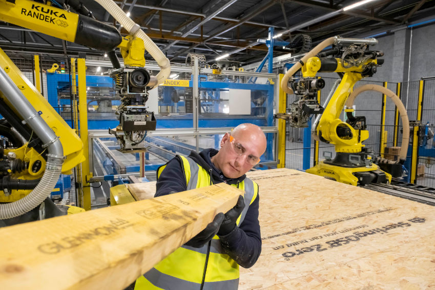 Man in a hi-vis vest holds a wood beam in a factory with yellow KUKA robots and plywood panels.