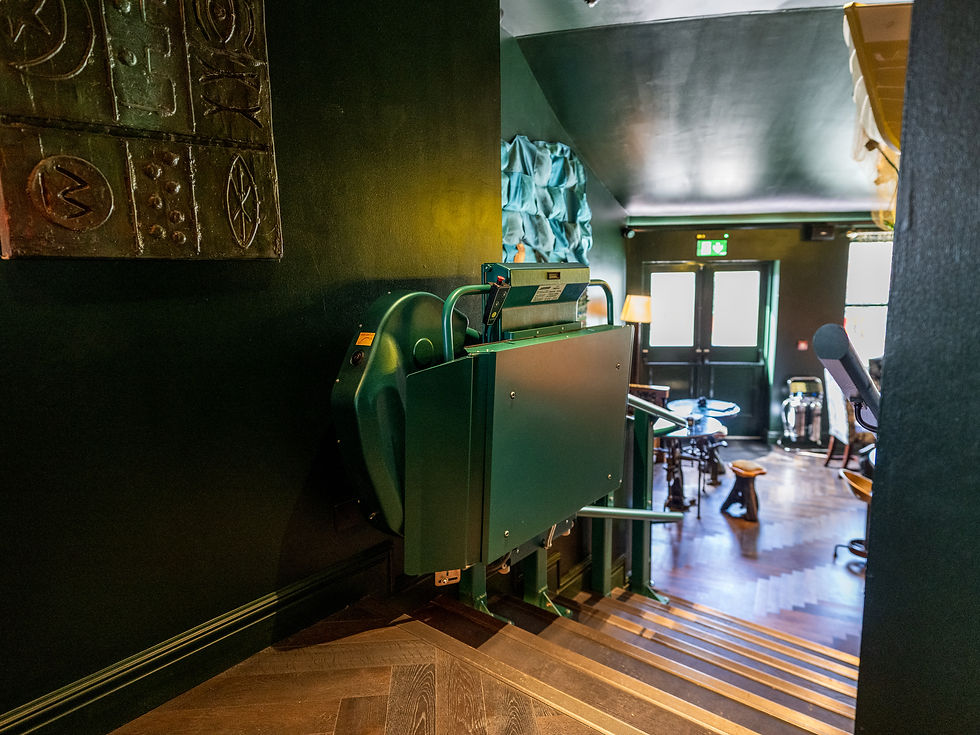 Green stairlift on dark stairs in a dimly lit room leading to a small, cozy cafe. Wooden tables and chairs are visible, with soft lighting.