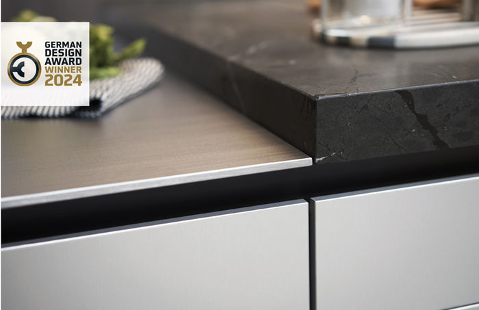 Modern kitchen counter with sleek steel and black marble surfaces. German Design Award Winner 2024 logo in upper left corner.