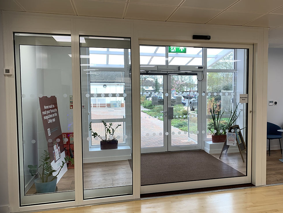 Glass entrance with plants and a poster inside. View of a garden and parked cars outside. Sign reads "Automatic door." Bright, inviting space.