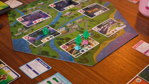 Plateau du jeu The Sims Board Game installé sur une table, avec cartes Lieux, pions Plumbob colorés et cartes d’action disposées autour.