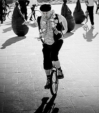 Black and white photo of Marty, a circus walkabout performer, balancing on his unicycle at Little Haven Beach in South Shields. He is dressed as a French mime character with a beret and a necklace of onions.