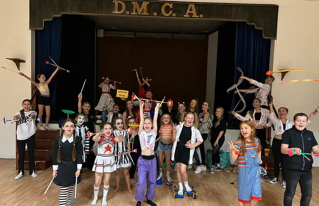 A large group of children and youth in Halloween costumes are celebrating after a circus skills workshop at the DMCA dance school party in Sunderland. They are holding props like devil sticks and balance boards.