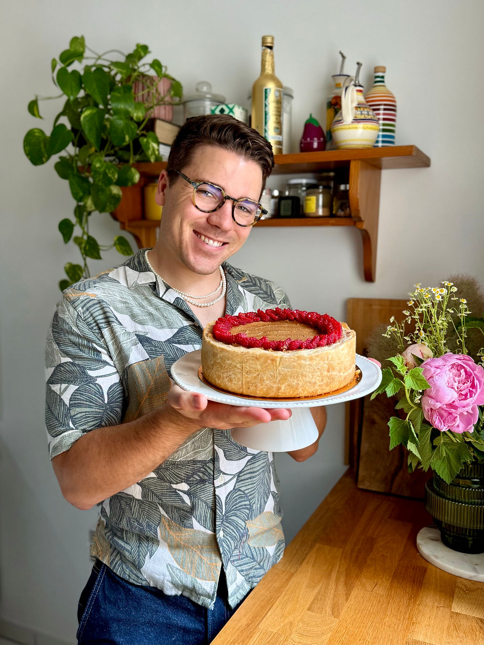 Homme souriant en chemise à motifs tenant un gâteau aux framboises dans une cuisine avec plantes et étagères. Flan Marbré Framboise Cacahuète.