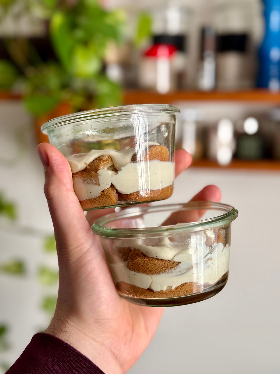 Deux verrines de tiramisu, crème et biscuits visibles. Arrière-plan flou avec étagères et plantes vertes. Ambiance gourmande.