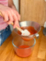 Personne verse un mélange rouge, utilisant une spatule et un tamis sur un bol en verre dans une cuisine en bois. Chemise rayée visible.