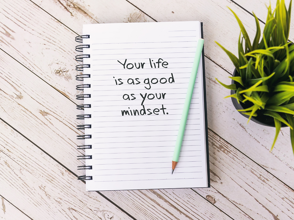 A spiral-bound note book open on a wooden table with the words "Your life is as good as your mindset" written on the open page. A green pencil lies on top of the notebook and a green house plants sits in a pot to the side of the notebook.