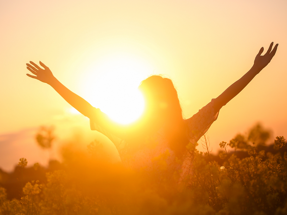 A woman standing in a field with tall flowers. The sun is setting in front of her and she is raising her arms to embrace the moment.
