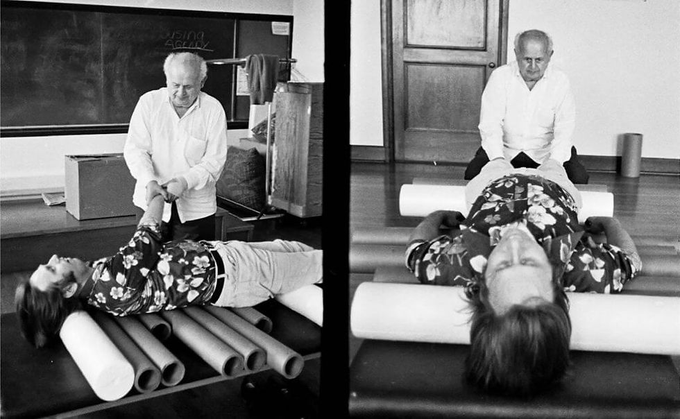 Moshe Feldenkrais pomáhá ležícímu muži na válcích. Černobílá fotografie, v pozadí tabule a dveře.