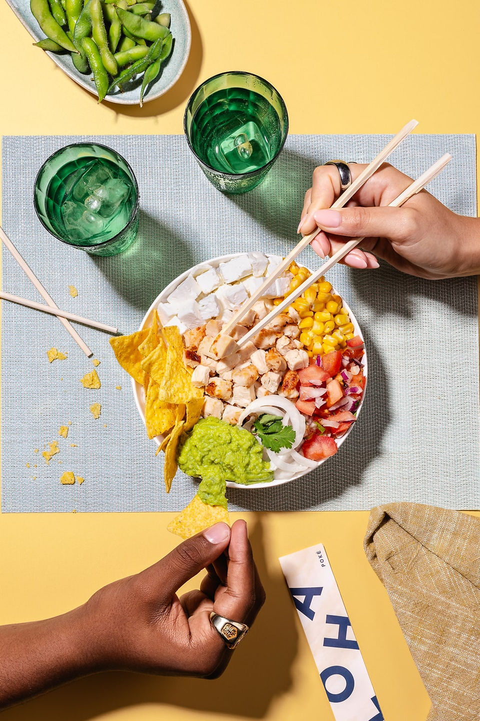Dos personas compartiendo un poke bowl con nachos, pollo, maíz, tomate y aguacate, usando palillos sobre una mesa amarilla con vasos verdes y edamame.