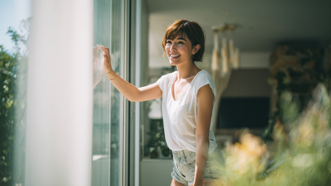 Smiling woman in white shirt and denim shorts cleans a glass door in a bright room, with greenery visible through the window. Relaxed mood.