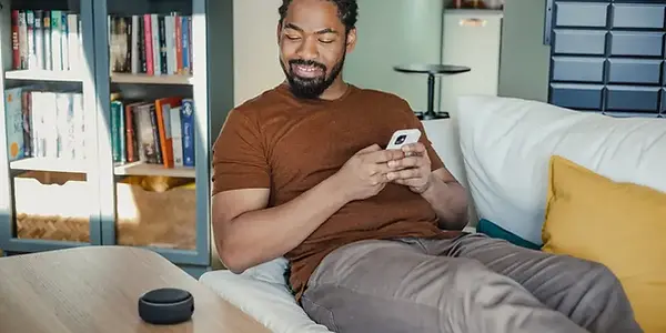 Bearded black man with smart phone on white couch smiling at Amazon Echo Dot