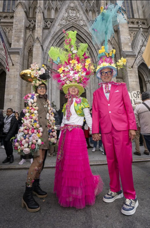 Colourfully dressed people wearing easter bonnets various designs with feathers, eggs, flowers, butterflies, ducks, rabbits in pink, yellow, white, light blue, light green and pink and lilac in front of a stone cathedral and a crowd gathered around on the pavement..