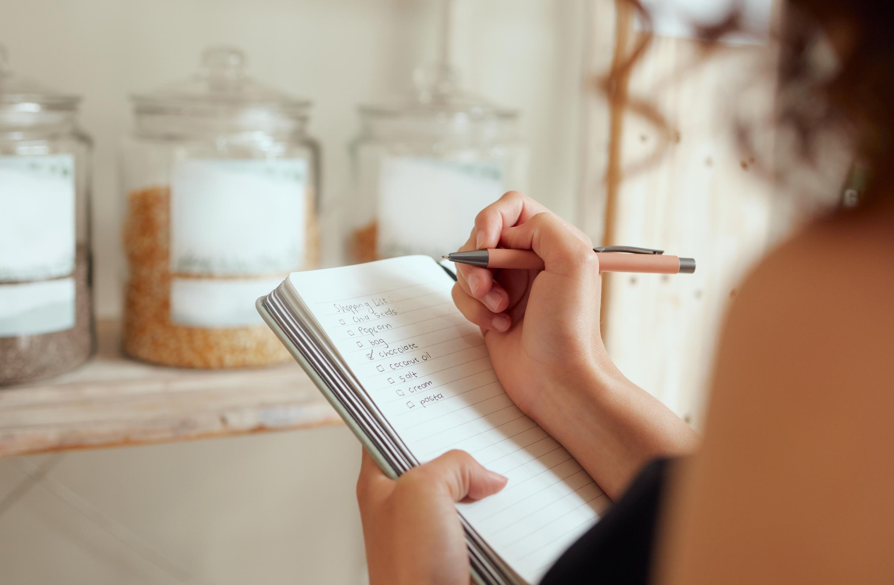 A person writing a list in a notebook beside jars of pantry staples