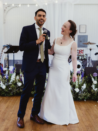 Ammar speaks into a microphone while Nancy smiles up at him during their wedding reception, standing in front of a live band setup and floral arrangements.