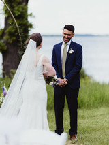 Ammar smiles warmly at Nancy during their outdoor wedding ceremony by the lake, with her veil flowing and bouquet in hand.