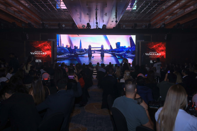 Audience seated in a corporate event, listening to a speaker on stage with large presentation screens displaying the event 