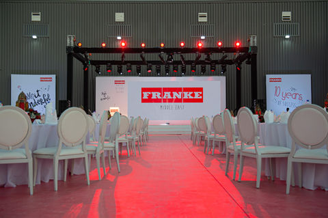 Wide view of the event setup, showcasing the stage with large screens and rows of tables arranged for attendees
