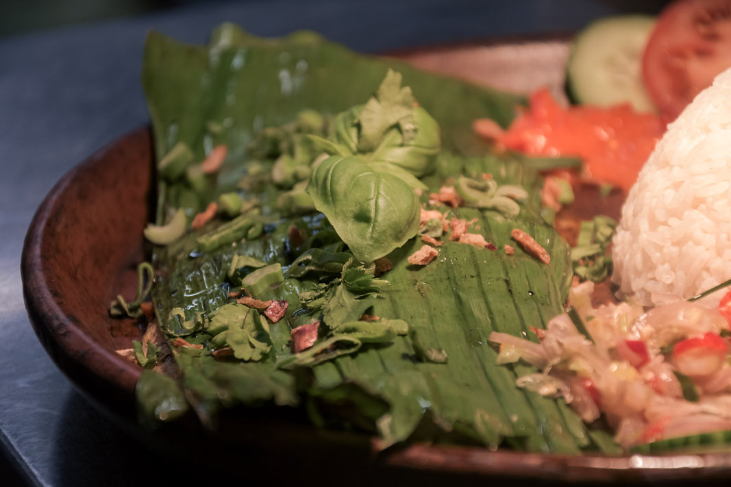 Grilled Indonesian Ikan Bakar wrapped in banana leaf, served with steamed rice, sambal, fresh herbs and sliced cucumber on a wooden plate at Bali Satay House.