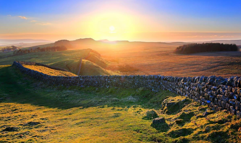 Sunset over a vast landscape with a stone wall and rolling hills. Golden light bathes the green and brown terrain, creating a serene mood. Countryside Walks