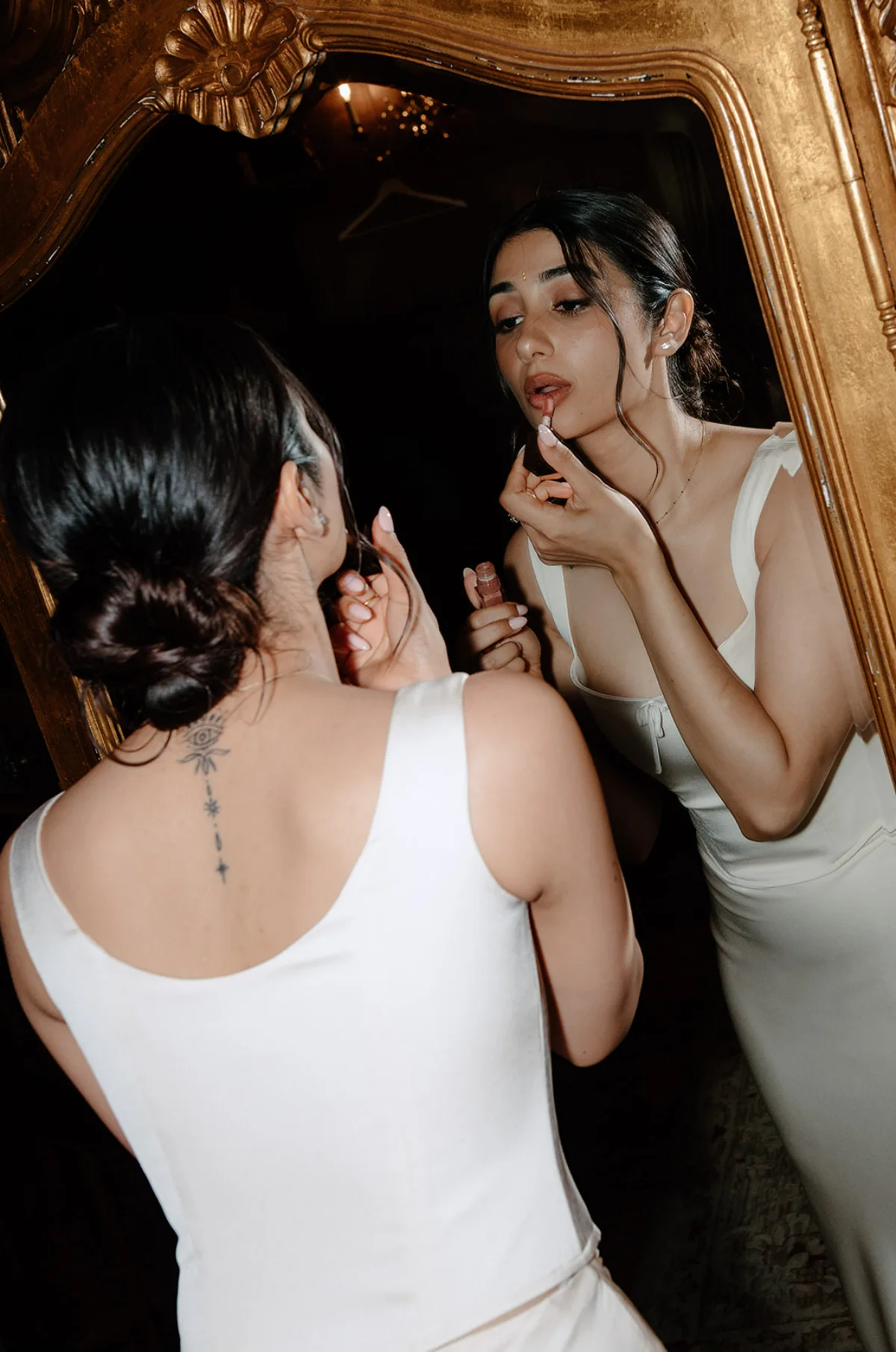 A bride wearing a sleek white gown puts lipstick on her lips in front of a mirror.