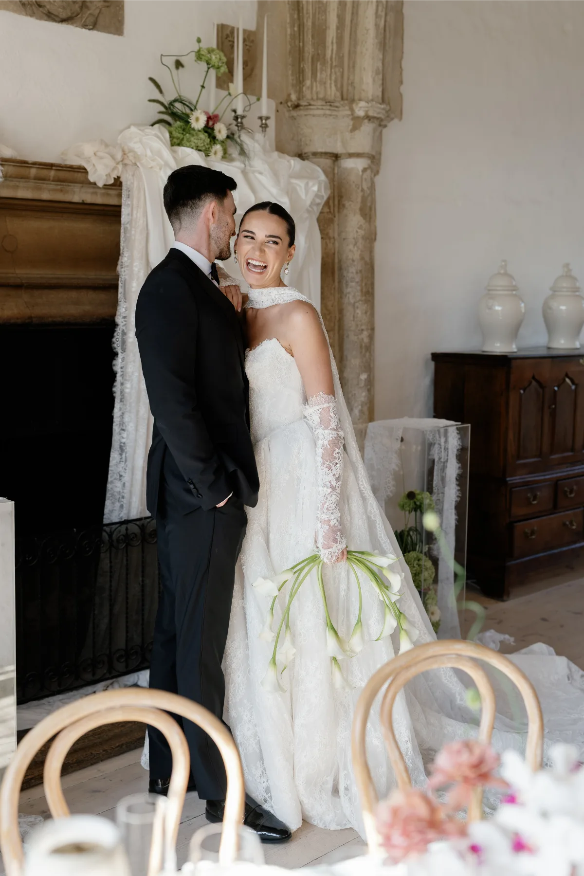 A bride wearing a wedding dress laughs with a man in a tuxedo as they stand together in front of an unlit fireplace, wearing makeup by Suffolk makeup artist Emily Chantal.