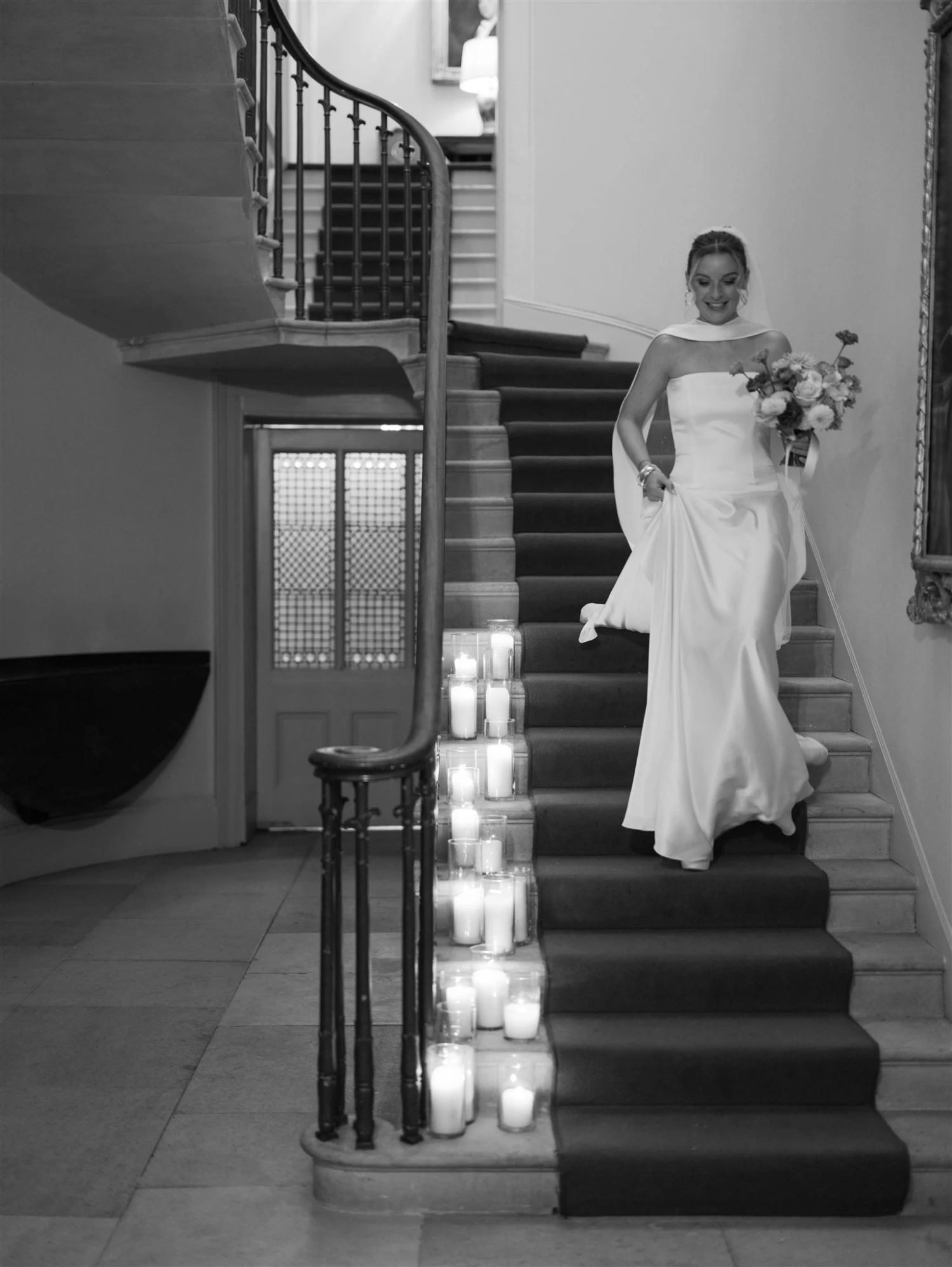 A bride walks down a grand staircase holding her dress in one hand and her bouquet in the other. Black and white image.