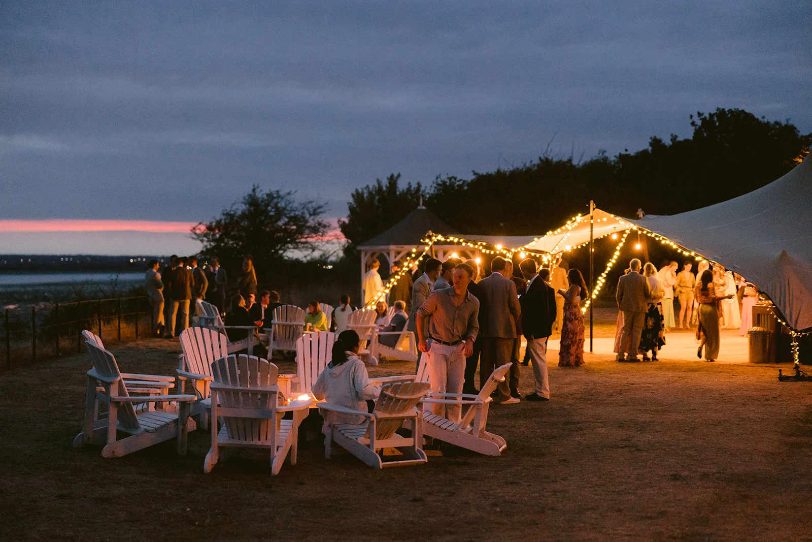 White deck chairs surround a firepit at sunset on Osea Island. Fairy lights glow in the distance as guests interact with each other.