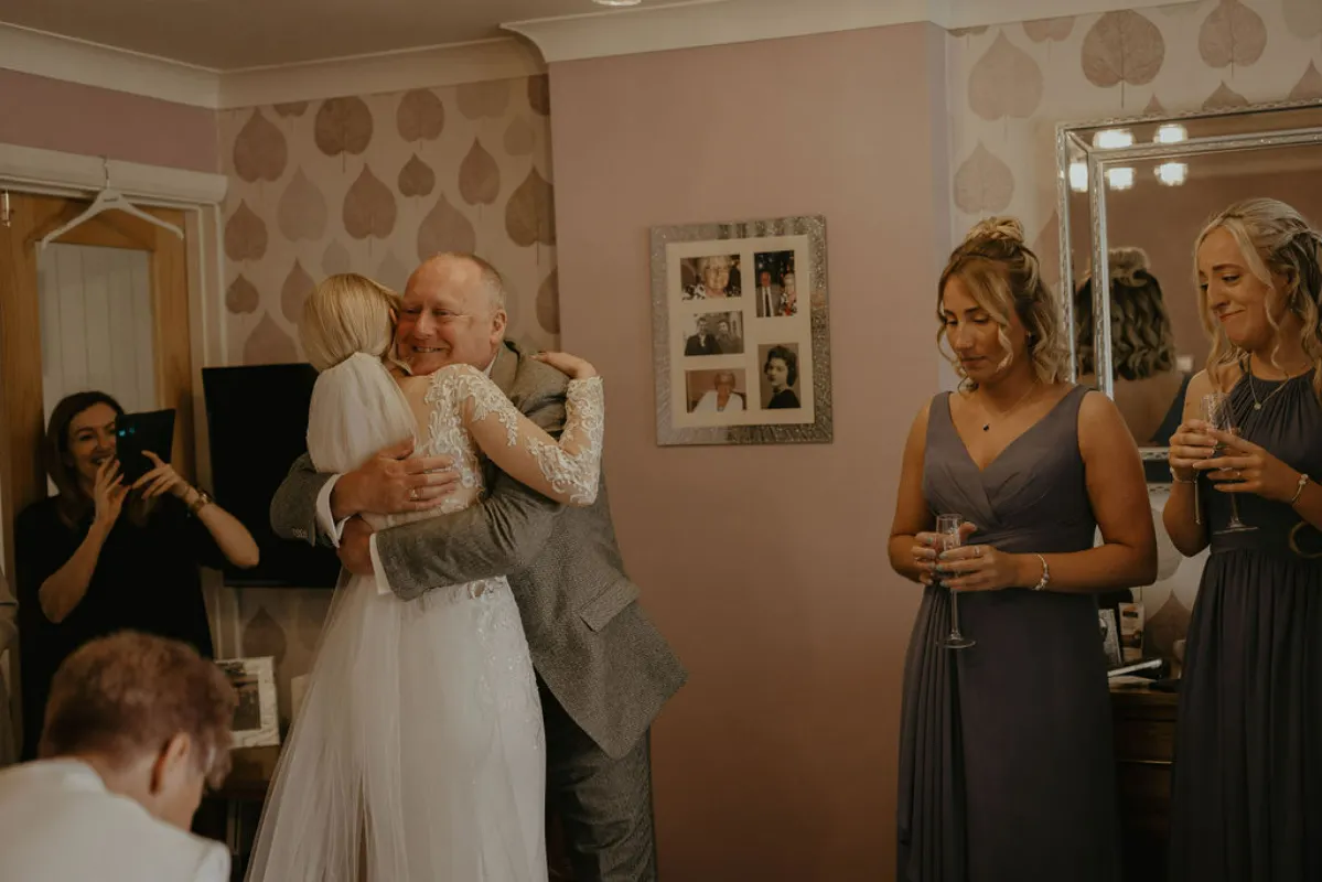 A bride hugs her father on a wedding day, surrounded by her bridal party. Suffolk wedding photography by Mikaela Jade.