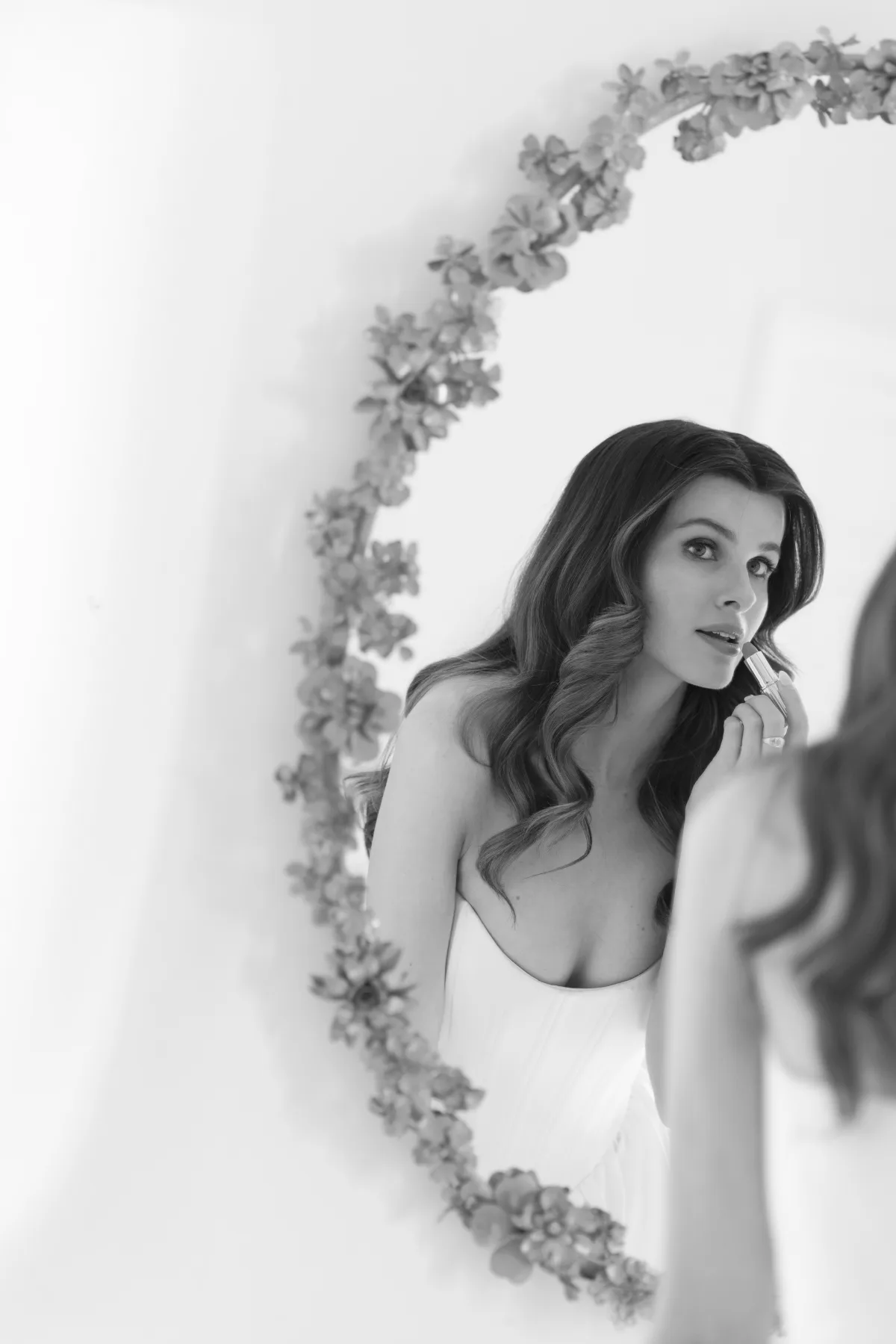 A bride applies lipstick in a mirror, wearing makeup by Suffolk bridal makeup artist Emily Chantal.