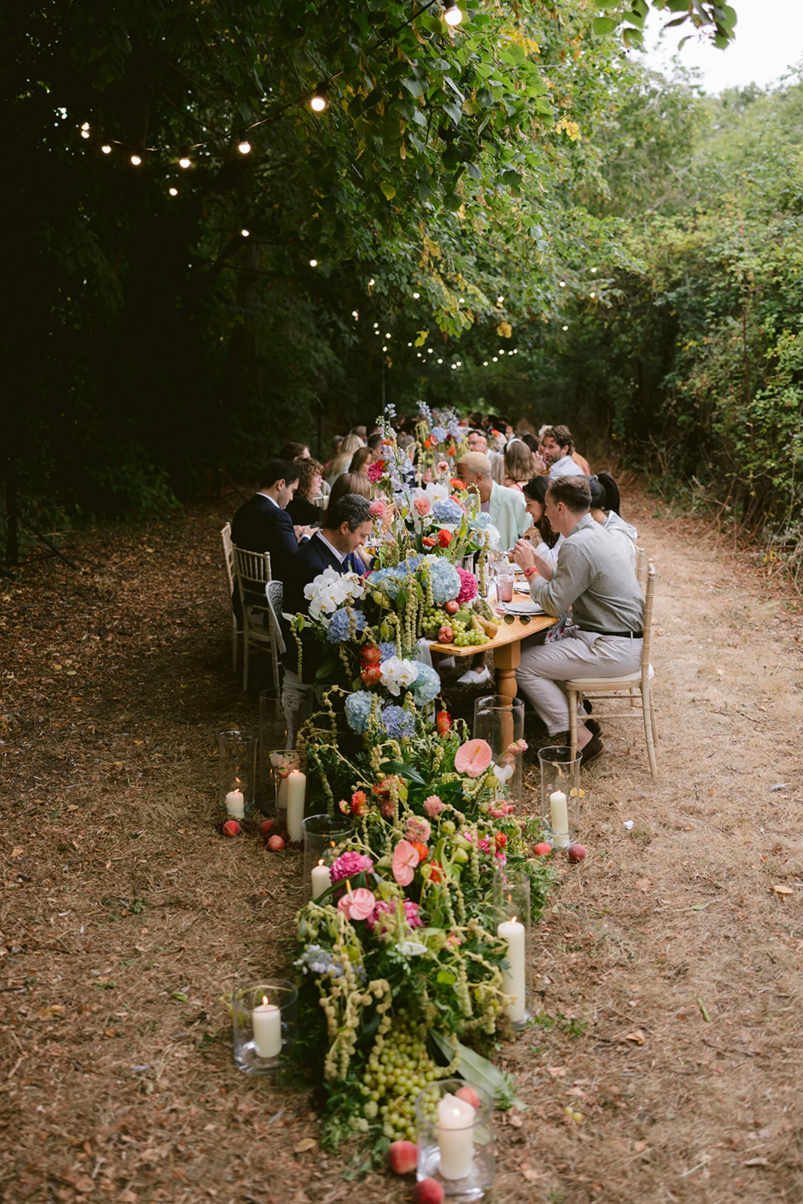 Guests eating and talking at a wedding breakfast table covered in colourful flowers, surrounded by trees, and with fairy lights hanging above.