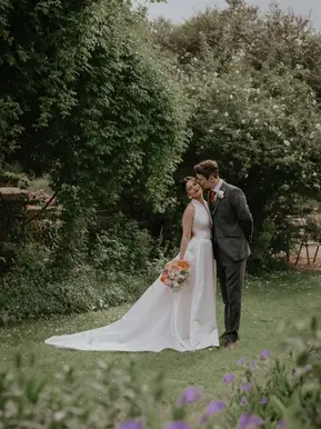 Bride in white gown holding flowers, groom in suit, smiling in garden. Greenery surrounds them with a brick building in the background. Photography by Mikaela Jade, Suffolk wedding photographer.