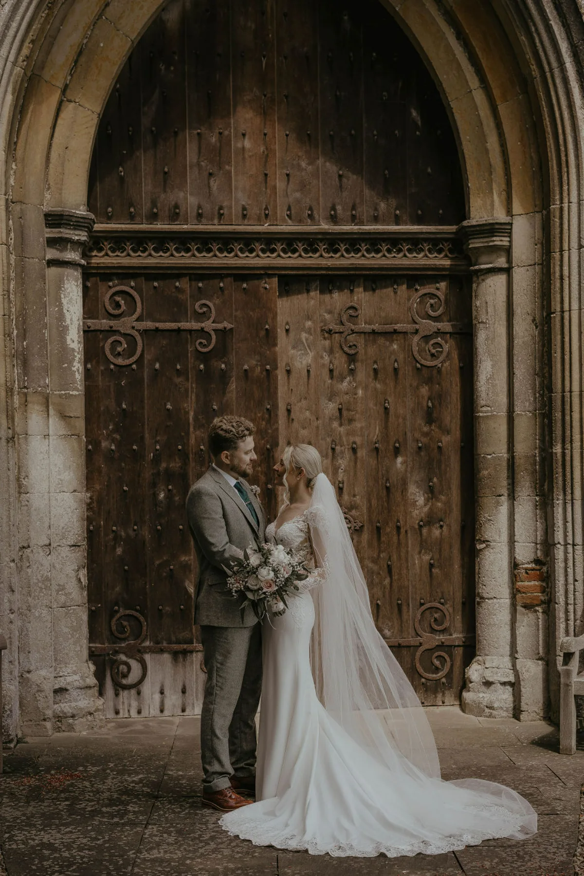 A bride and groom look into each other's eyes in front of a large, wooden church door. Suffolk wedding photography by Mikaela Jade.