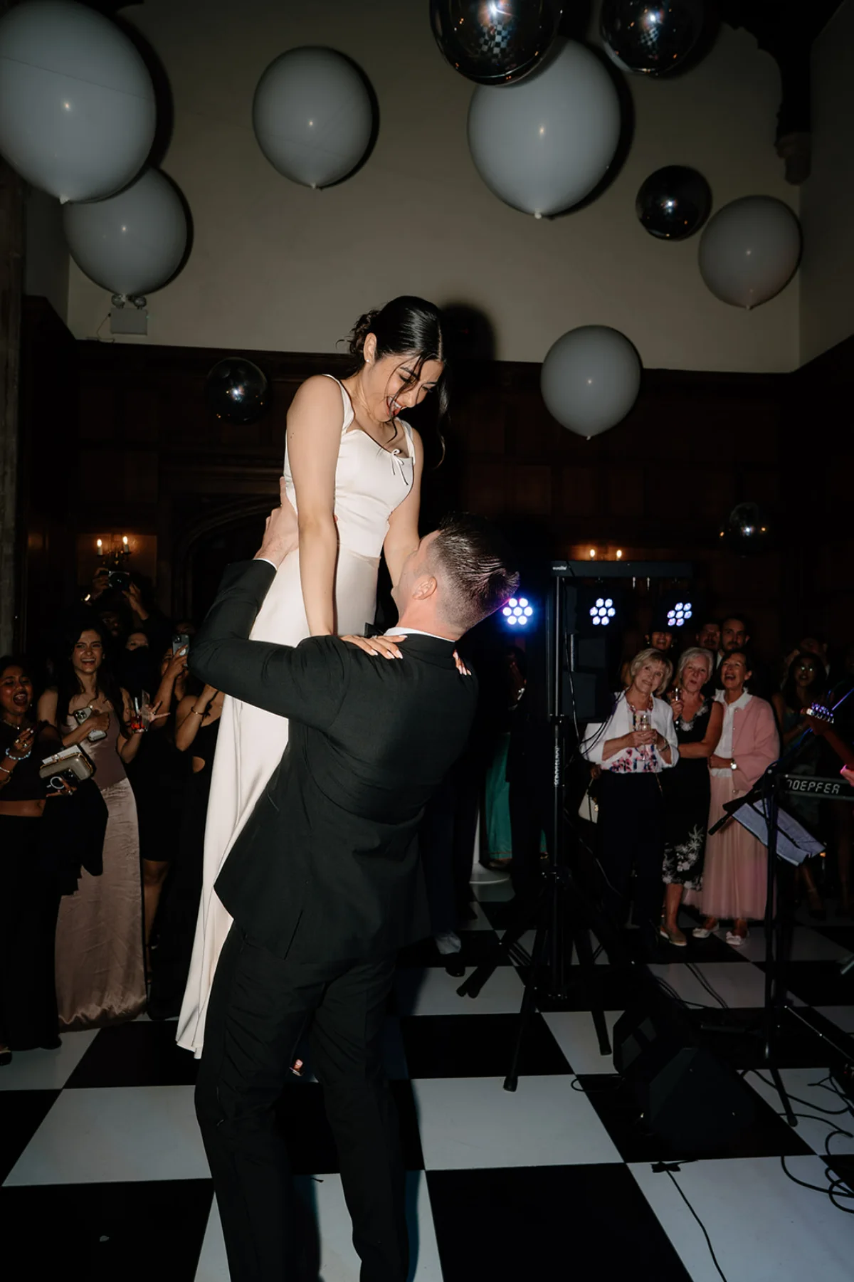 A groom lifts his bride into the air at their wedding party in Hengrave Hall, Suffolk, surrounded by guests and white balloons.