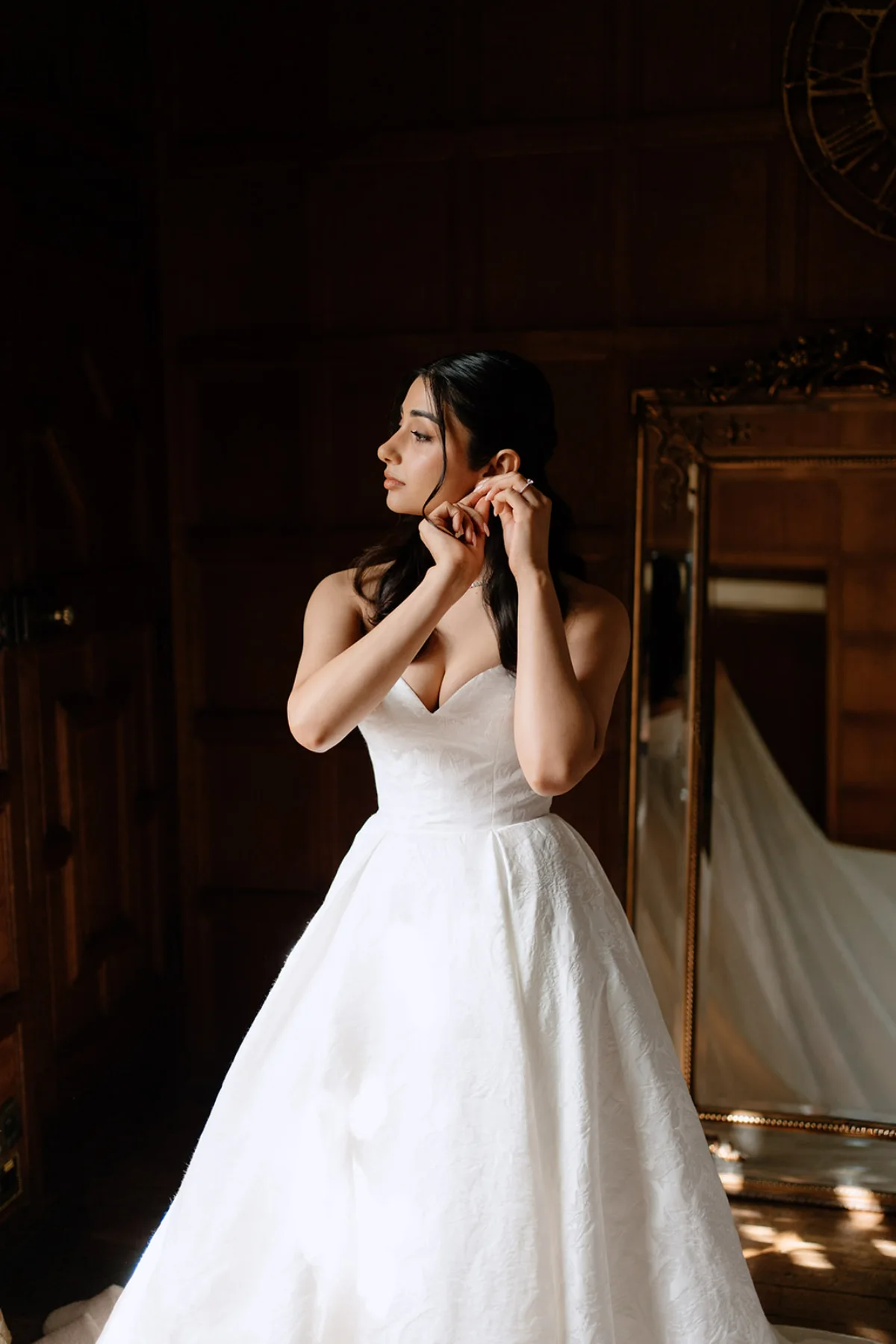 A bride puts on her earrings while wearing a white dress, looking to her side.