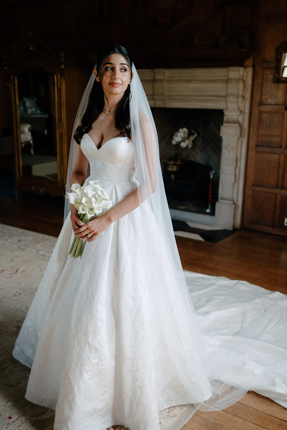A beautiful bride poses with a bouquet of white calla lilies in her long, flowing dress and long veil, while looking away into the light. Makeup by Emily Chantal.